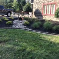 A landscaped front yard features a decorative stone dry creek bed, garden beds with ornamental grasses, and a brick house.