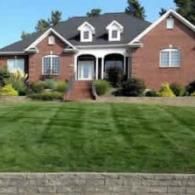 A two-story red brick house with a dark roof and manicured green lawn in front, viewed from a slightly low angle.