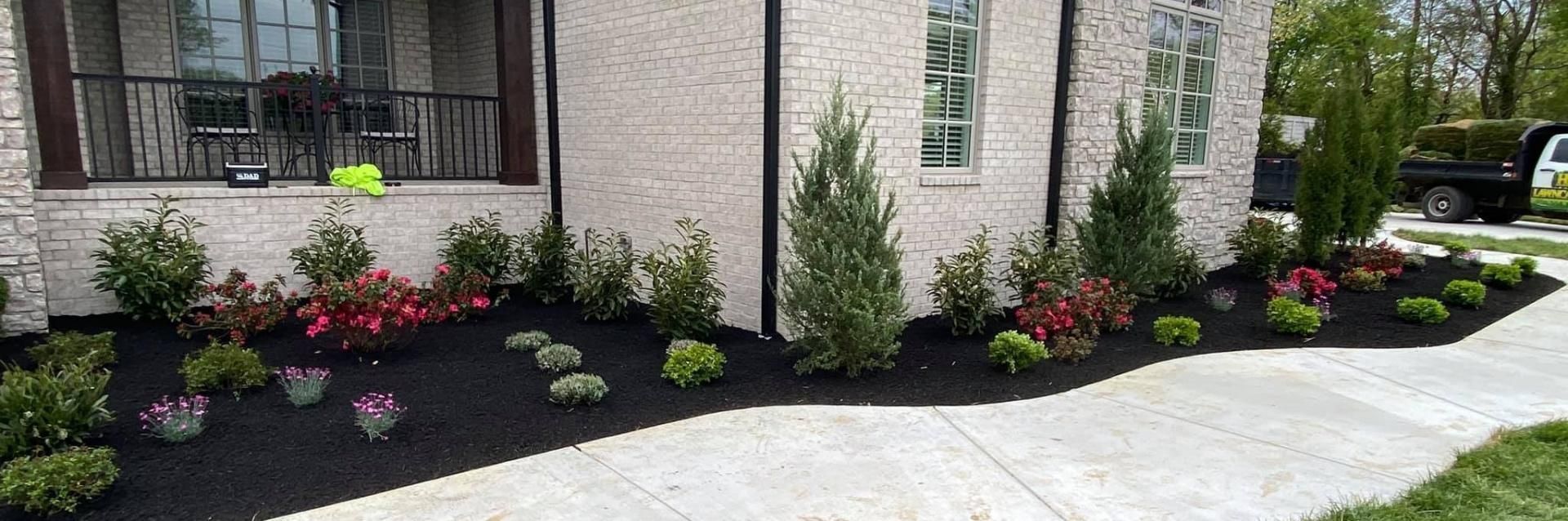 Landscaped garden bed with dark mulch, various green shrubs, and colorful flowers in front of a stone-faced house.