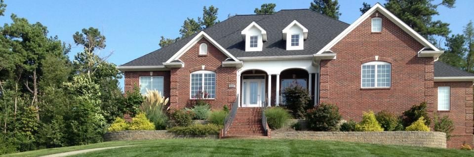 A two-story brick house with a dark shingled roof, arched windows, and a landscaped front yard with a low stone wall.