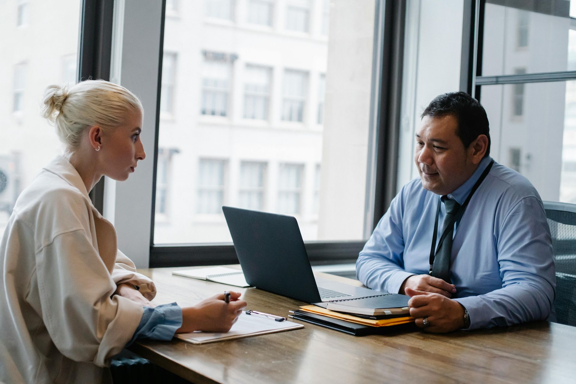 Two professionals sitting across from each other at a wooden desk with a laptop, engaged in a conversation in an office.