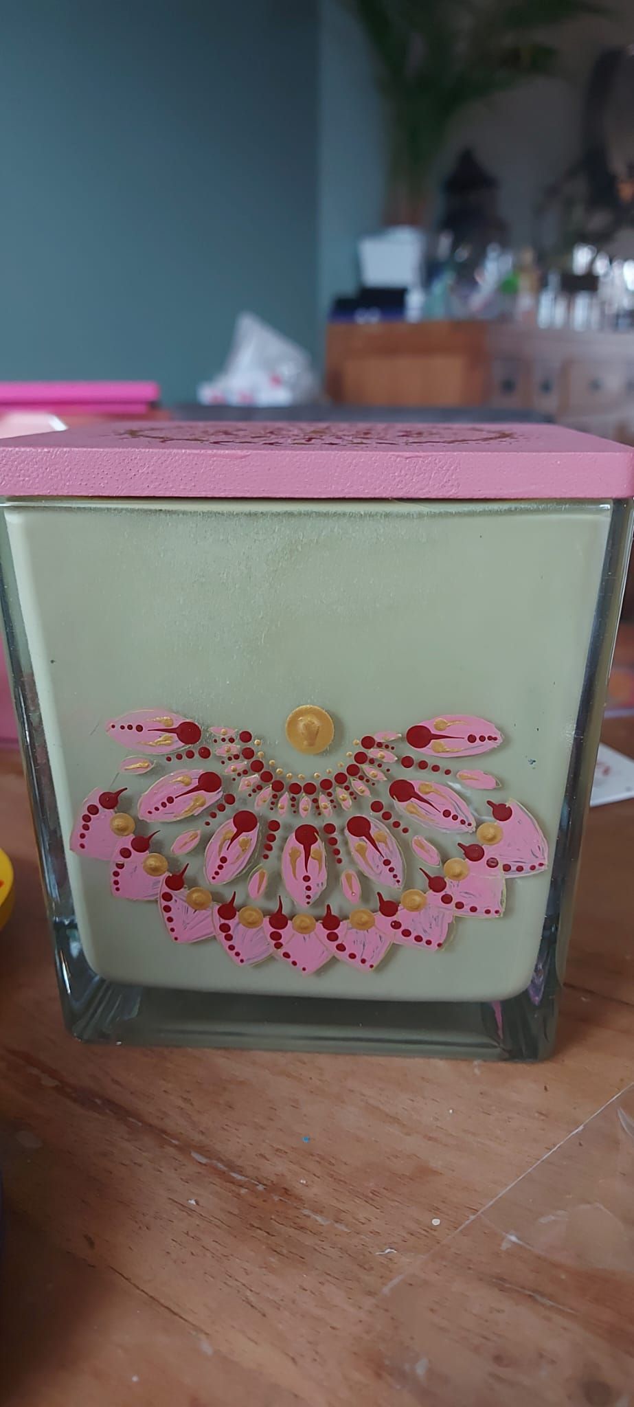 Een decoratieve kaars in een vierkante glazen houder, met een roze deksel en een roze en gouden bloemmotief aan de voorkant.