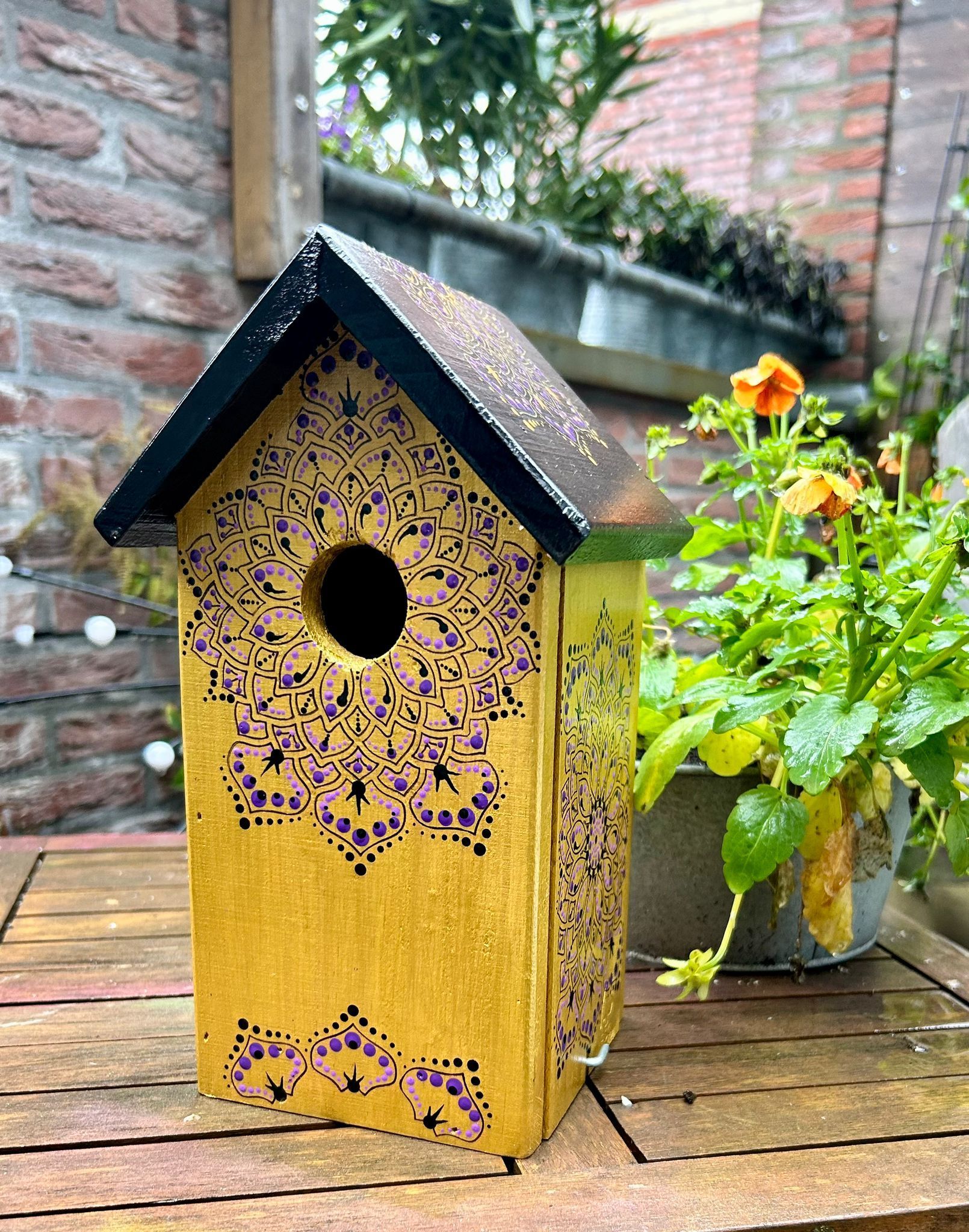 Geel vogelhuisje met zwart dak en paars mandala-motief, op een houten tafel buiten.
