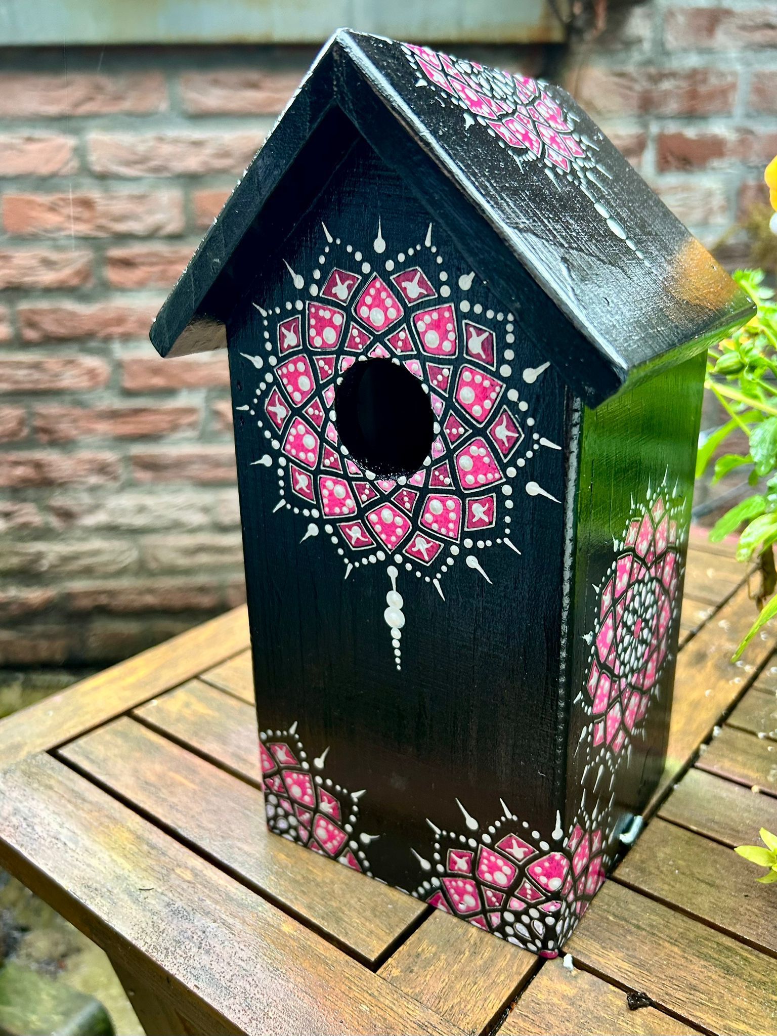 Een zwart vogelhuisje, versierd met roze en witte mandala-motieven, staat op een houten tafel.