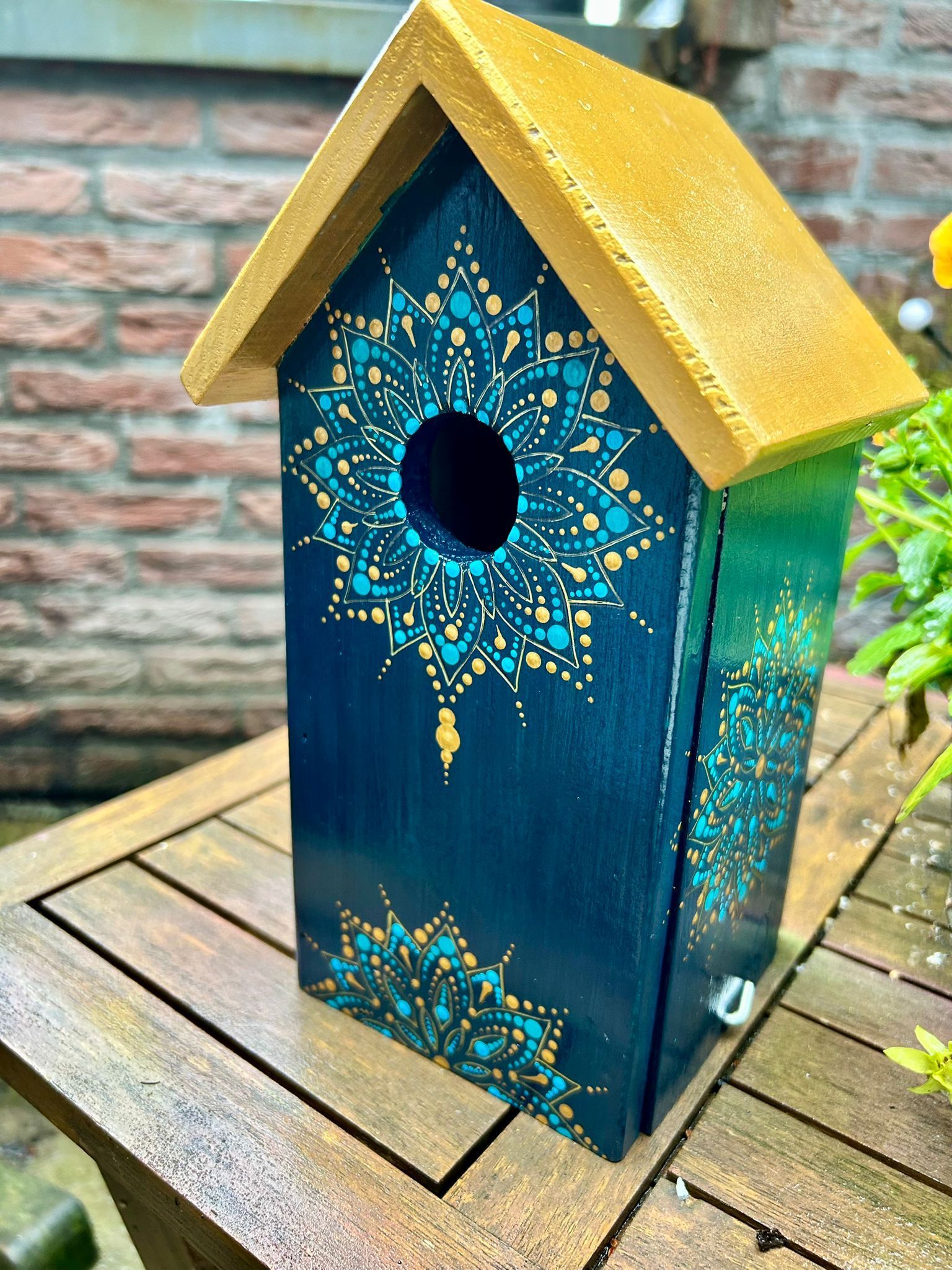 Donkerblauw vogelhuisje met turquoise en gouden mandala-motieven, gouden dak, op een houten tafel.