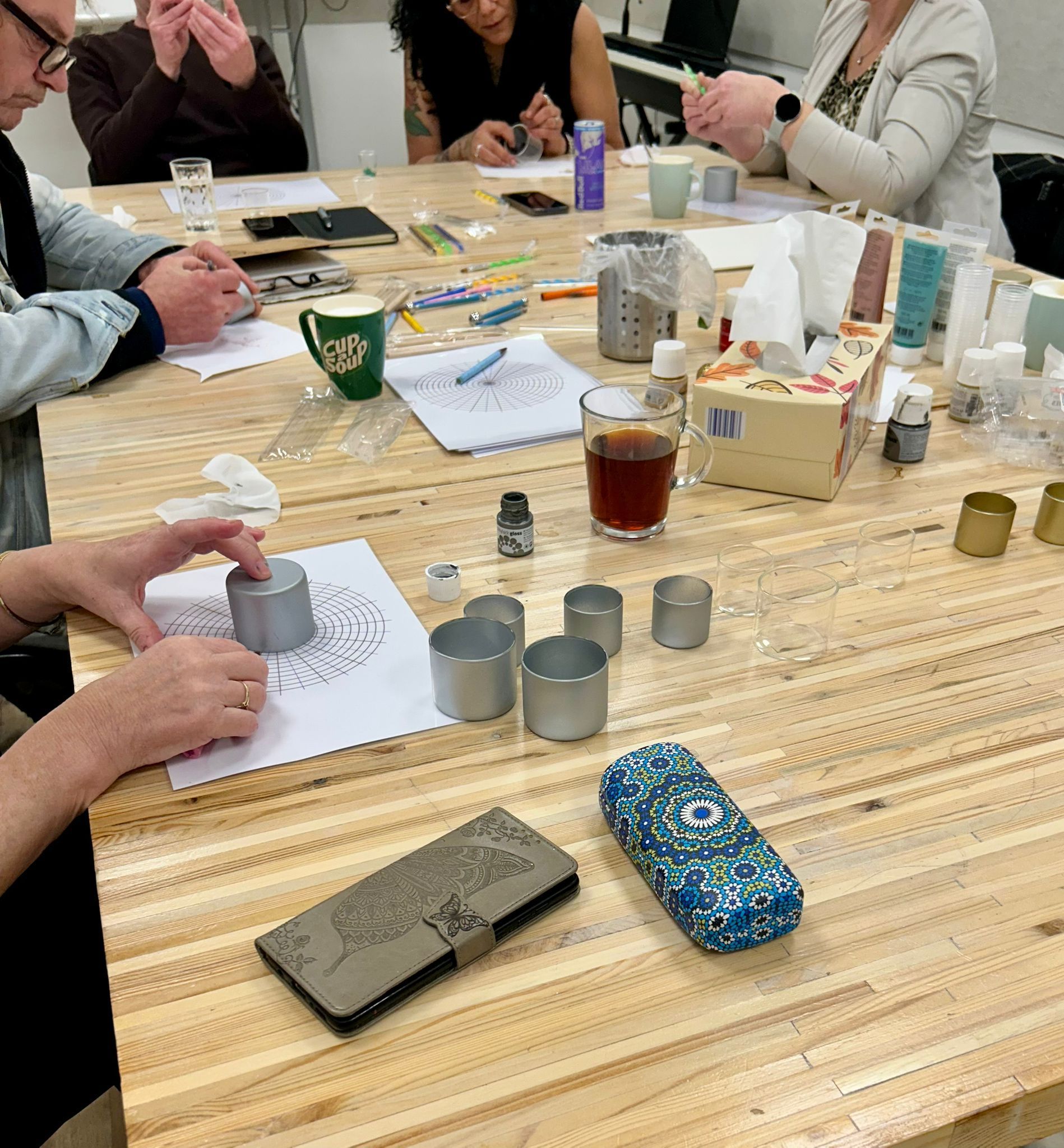Mensen zitten aan een tafel kaarsen te maken. Benodigdheden en aantekeningen liggen verspreid over het houten tafelblad.