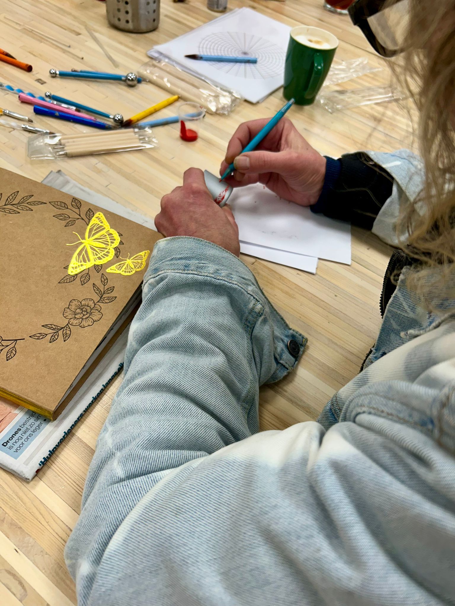 Een persoon tekent met een pen aan een tafel. Een notitieboekje met gouden accenten en tekenbenodigdheden liggen in de buurt.