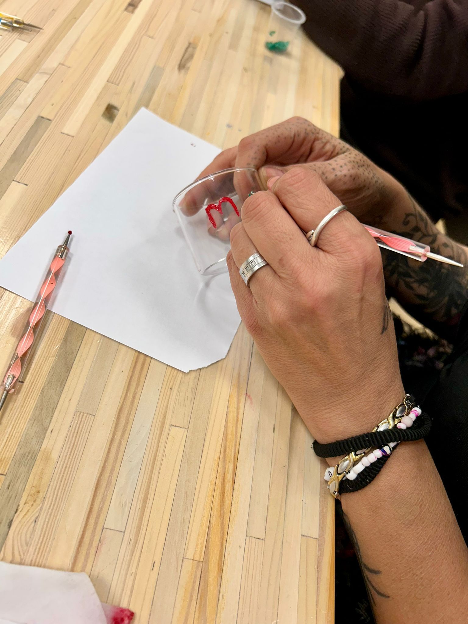 Een persoon die aan een houten tafel zit en een rode letter op een glas schildert.