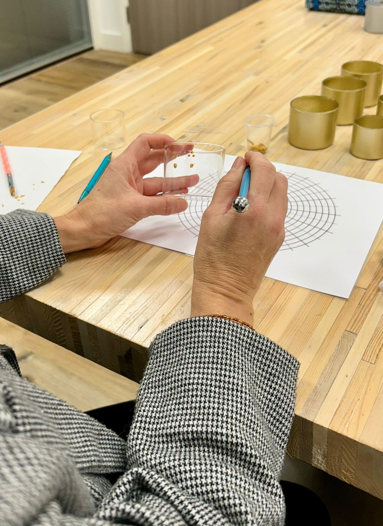 Een persoon gebruikt een potlood om een cirkelvormig ontwerp in te kleuren; gouden bakjes staan op tafel.