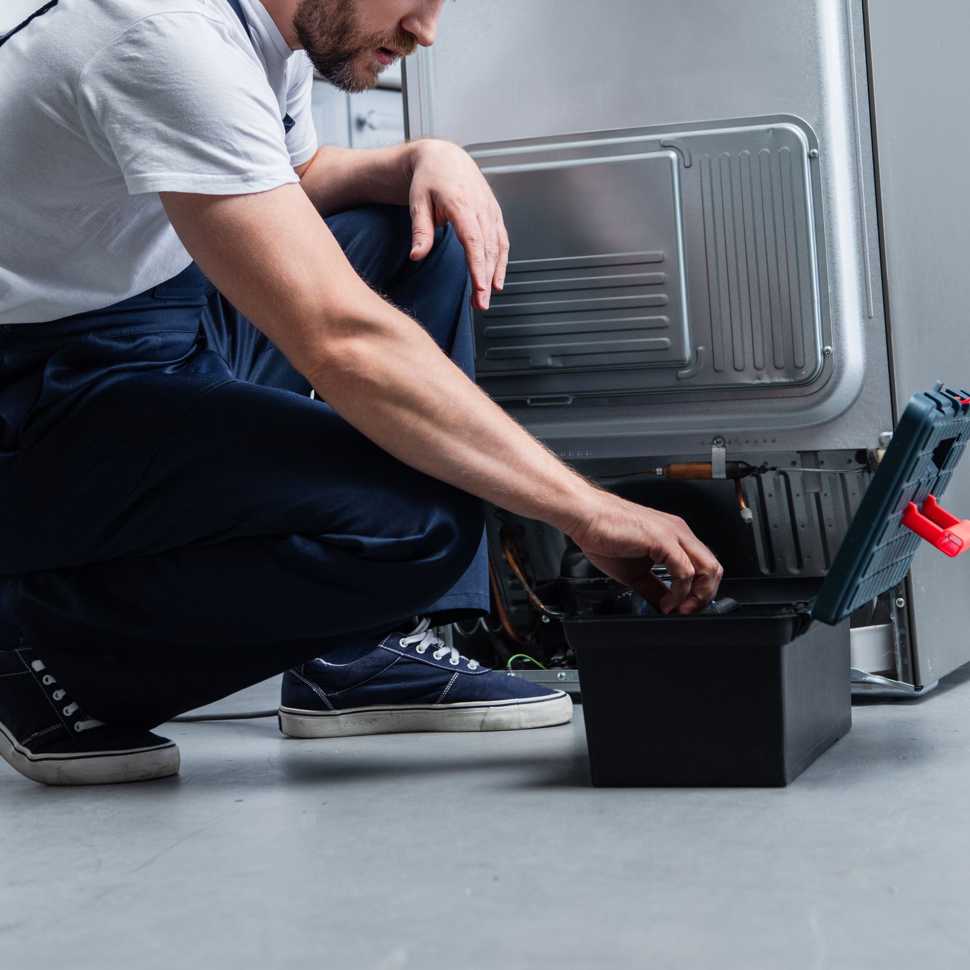 A man is kneeling down to fix a refrigerator.