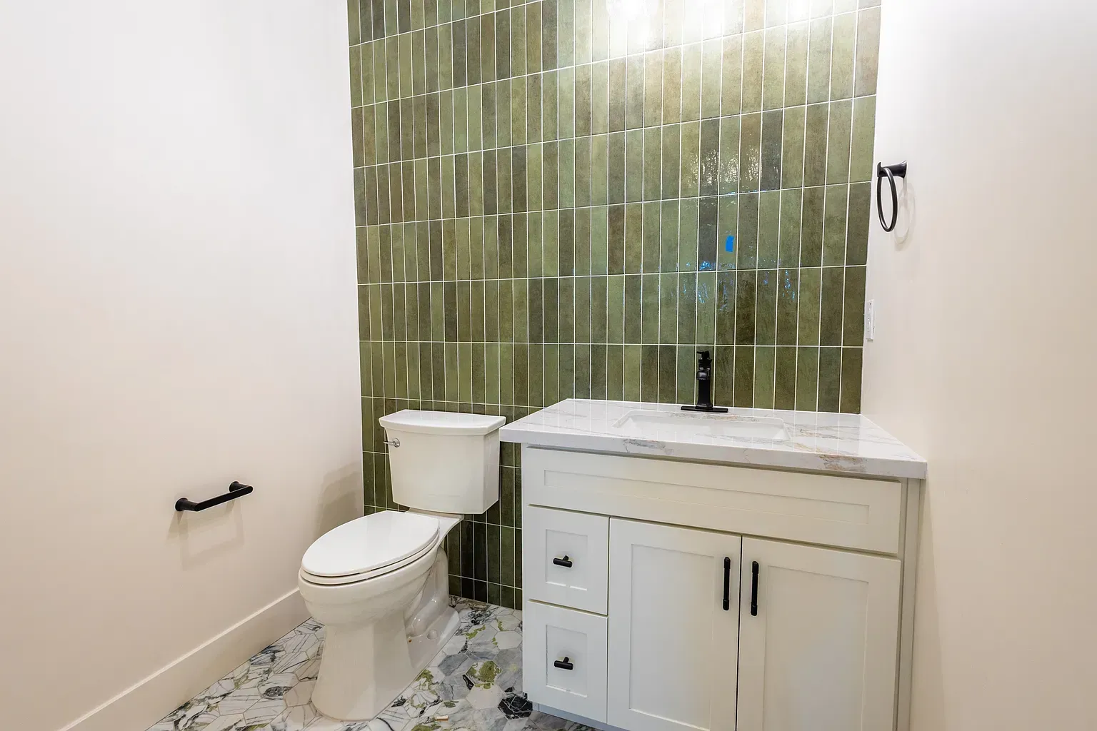 A powder room with a white vanity and toilet. Green tiled accent wall. Floral floor tiles.