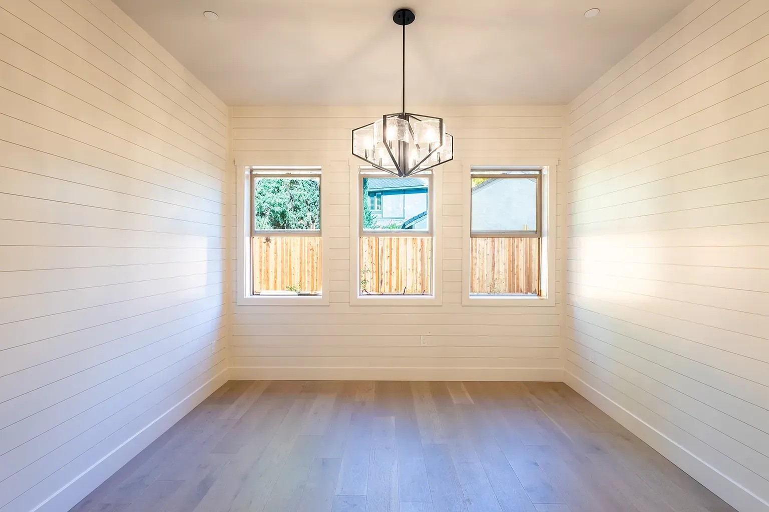 Empty room with three windows and a chandelier. Light wood floors and shiplap walls.