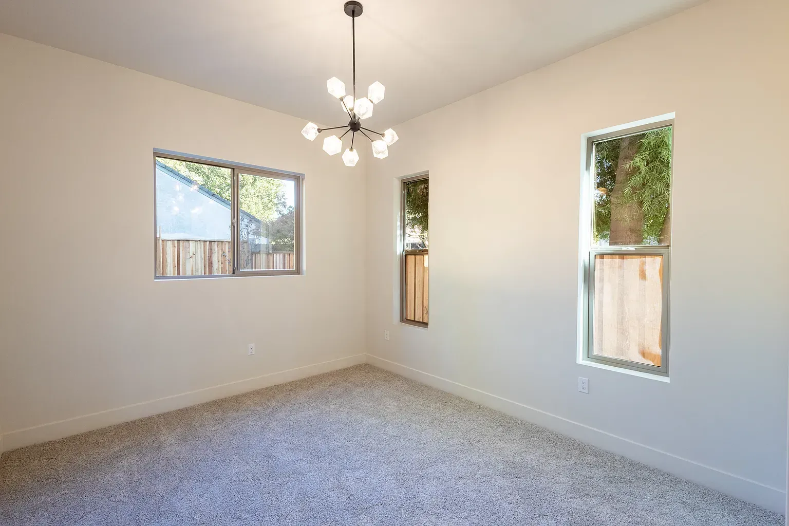Empty room with three windows, beige walls, and light fixture, carpeted floor.