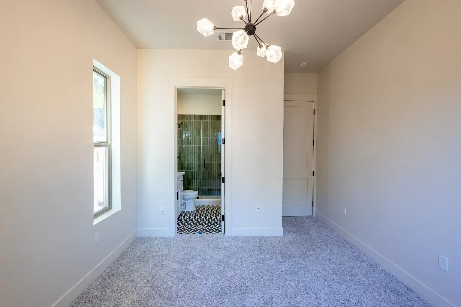 Empty bedroom with bathroom access, light walls, patterned carpet, and chandelier.