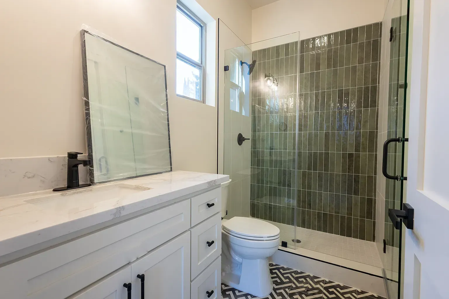 Bathroom with white vanity, patterned floor, green tiled shower, and a toilet.