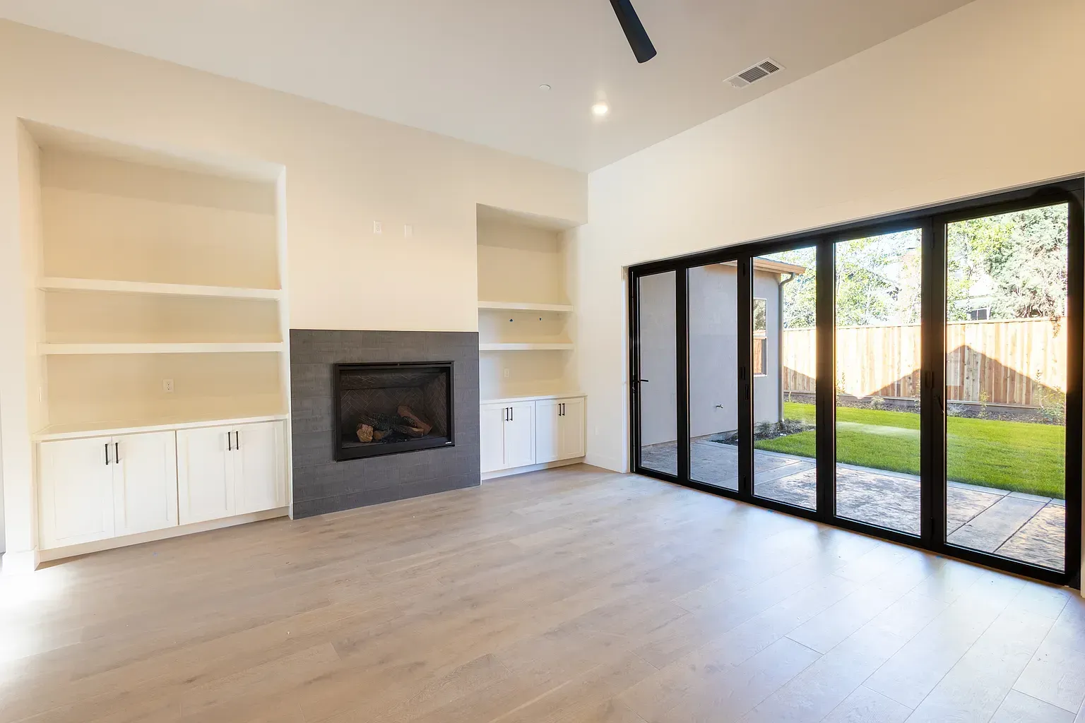 Empty modern living room with fireplace, built-in shelves, and large sliding glass doors to a backyard.