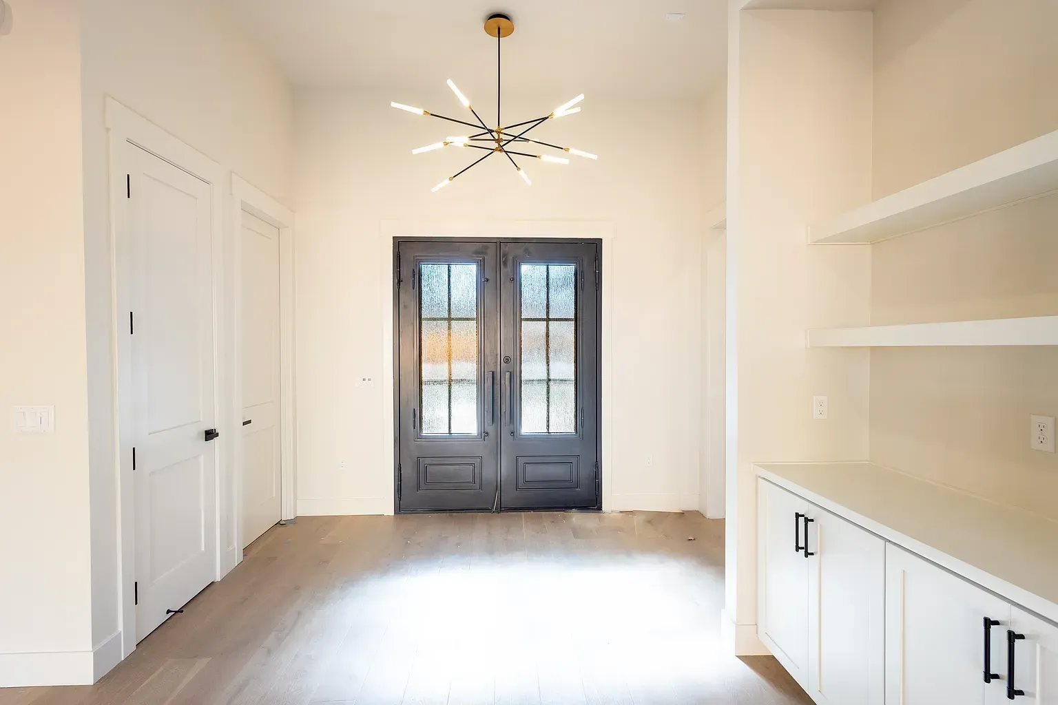 Hallway with white walls, wood floor, double doors, and modern chandelier.