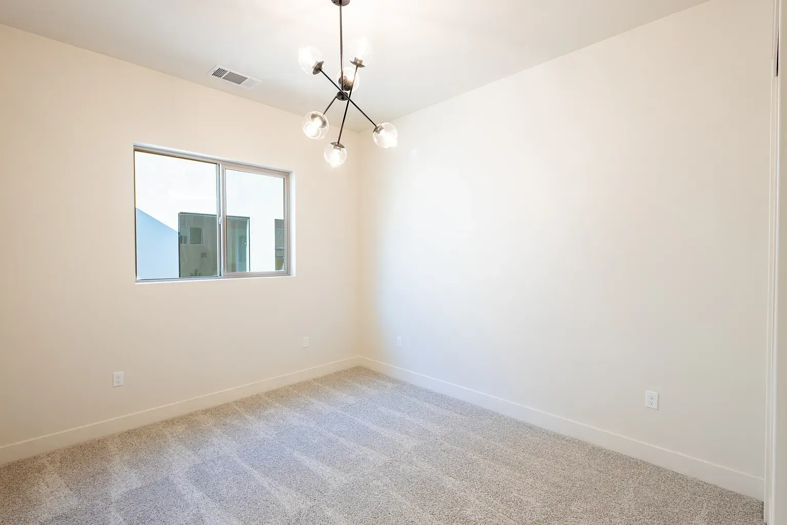 Empty room with beige walls, window, carpet, and modern chandelier.