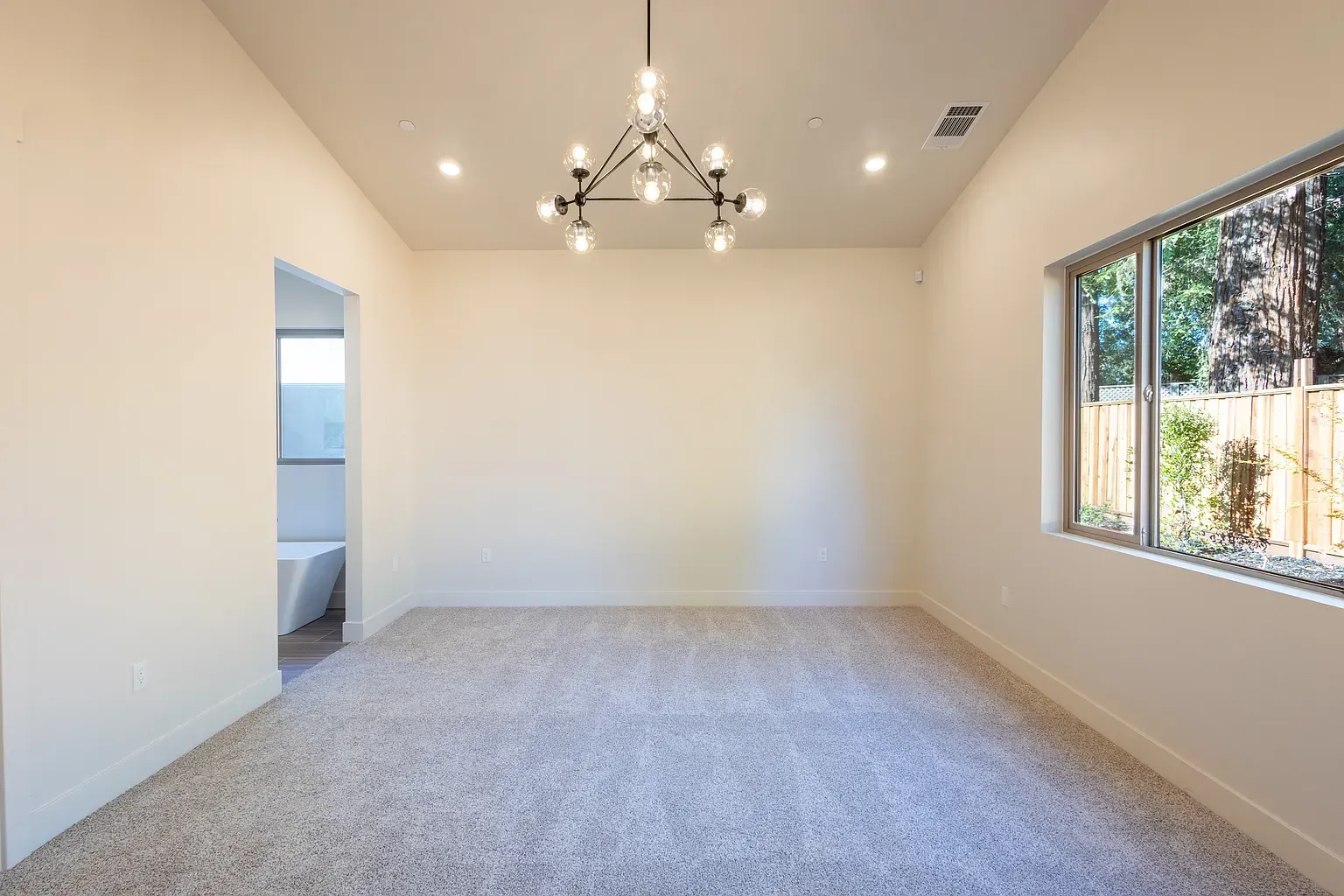 Empty room with light carpet, a window, and a modern chandelier. Doorway to another room on the left.