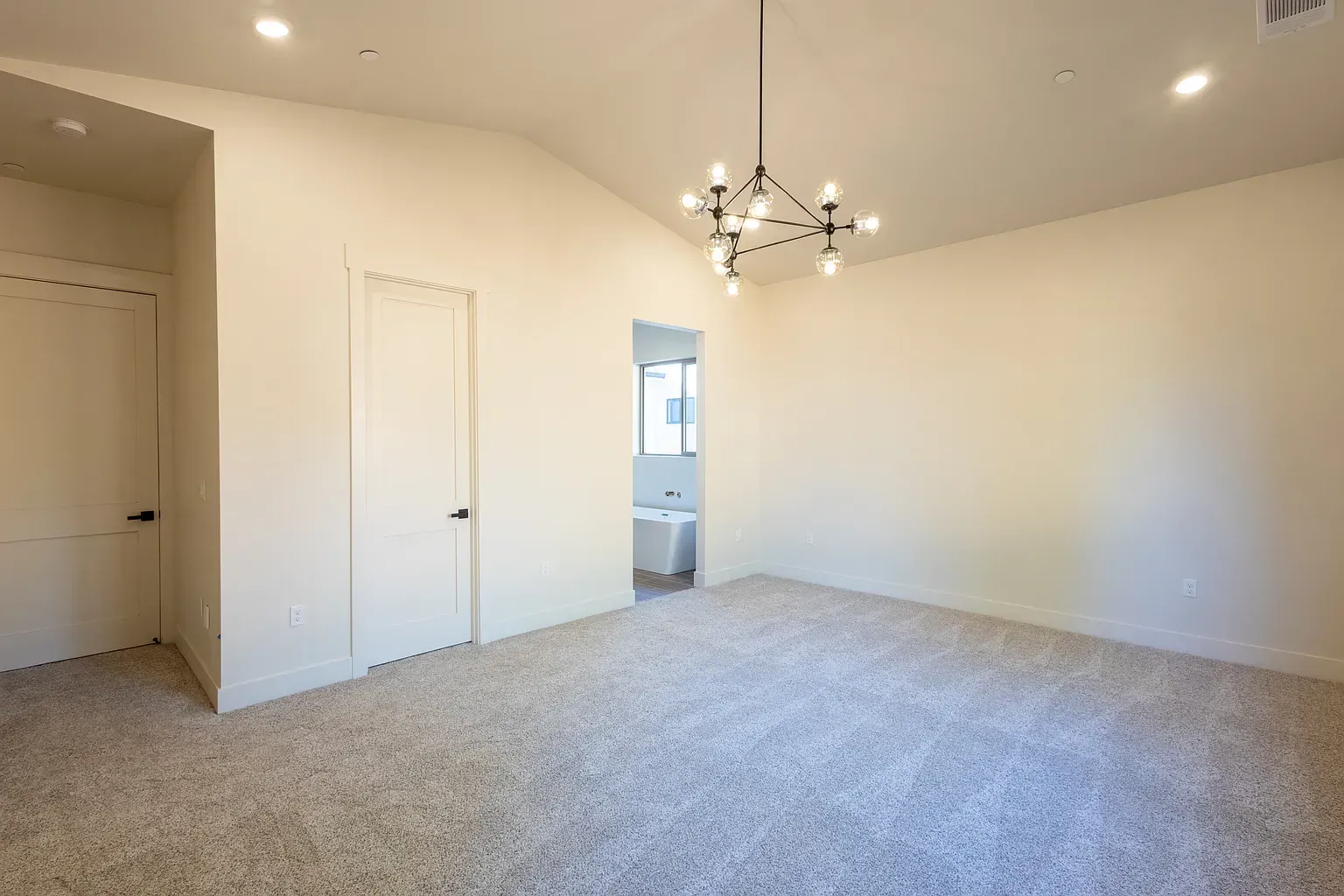 Empty room with beige walls, light-colored carpet, and a chandelier. A doorway leads to a bathroom.