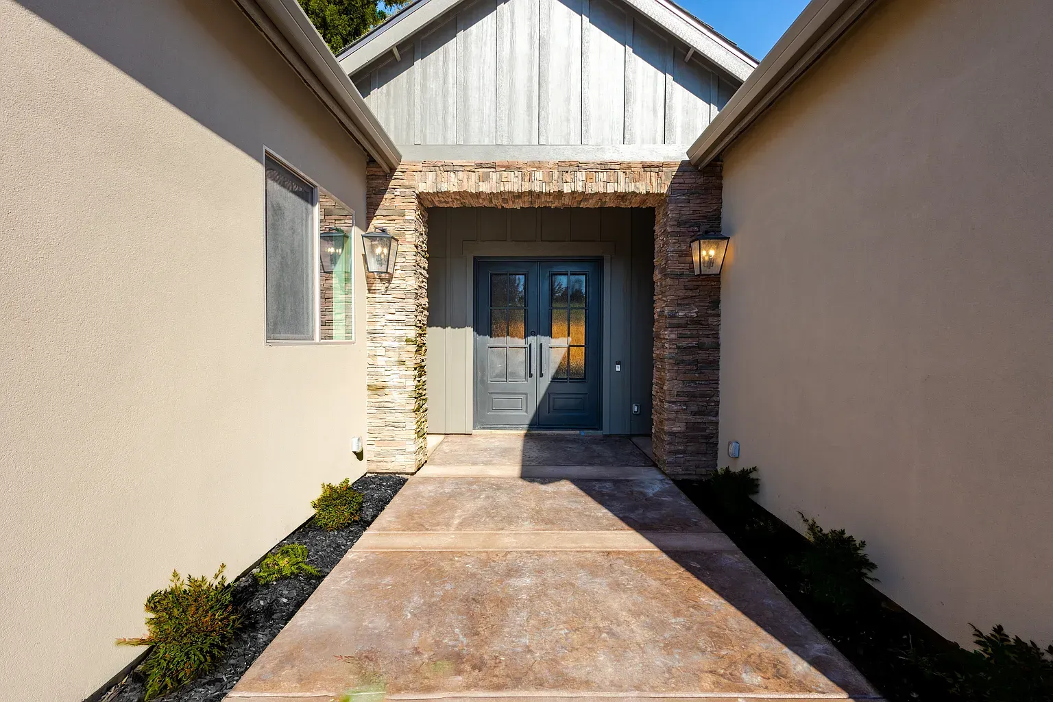 Exterior view of a home's entrance: stone columns frame a gray door, leading to a concrete path.