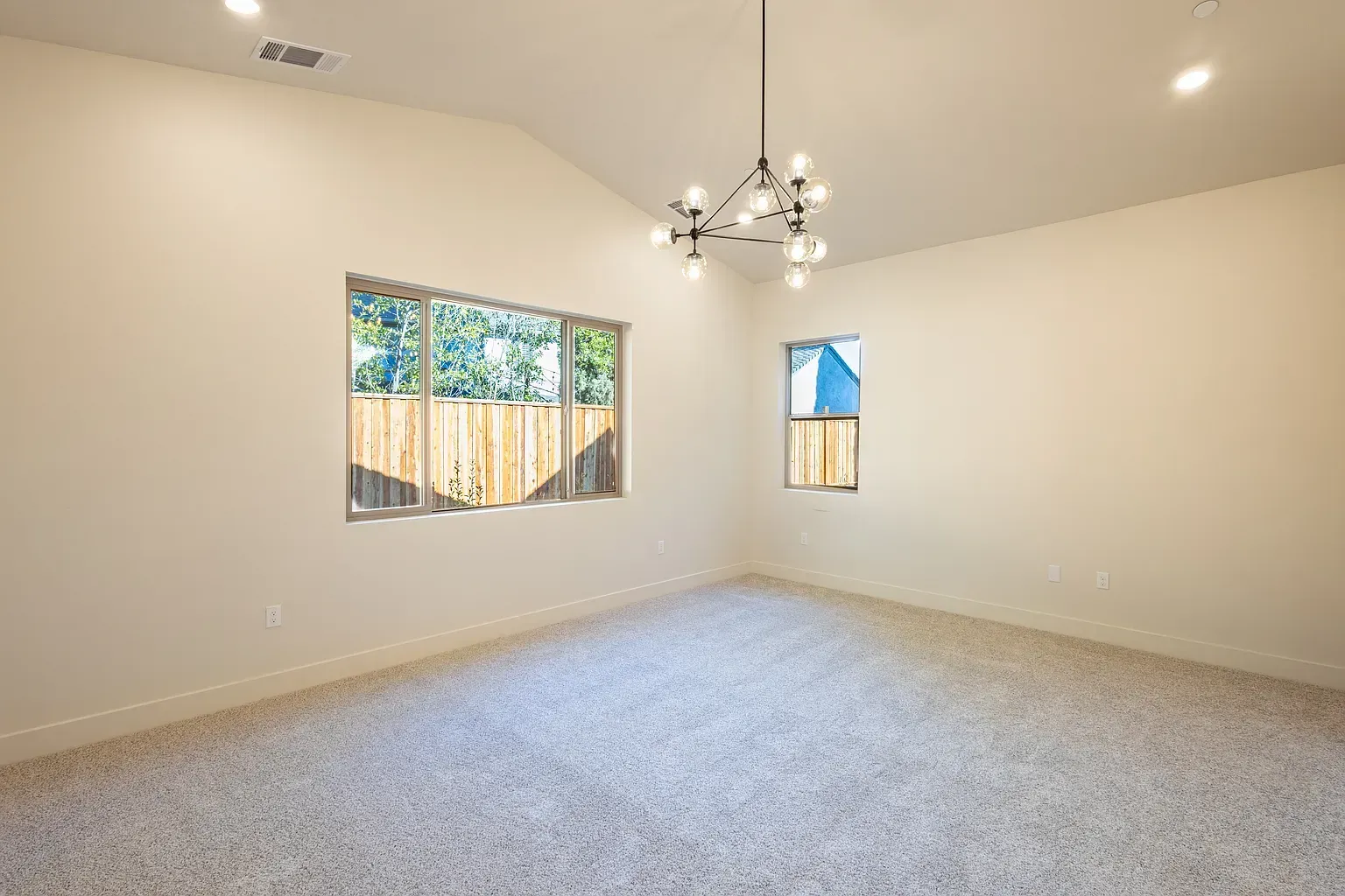 Empty room with beige walls, carpet, and a modern chandelier. Two windows provide natural light.