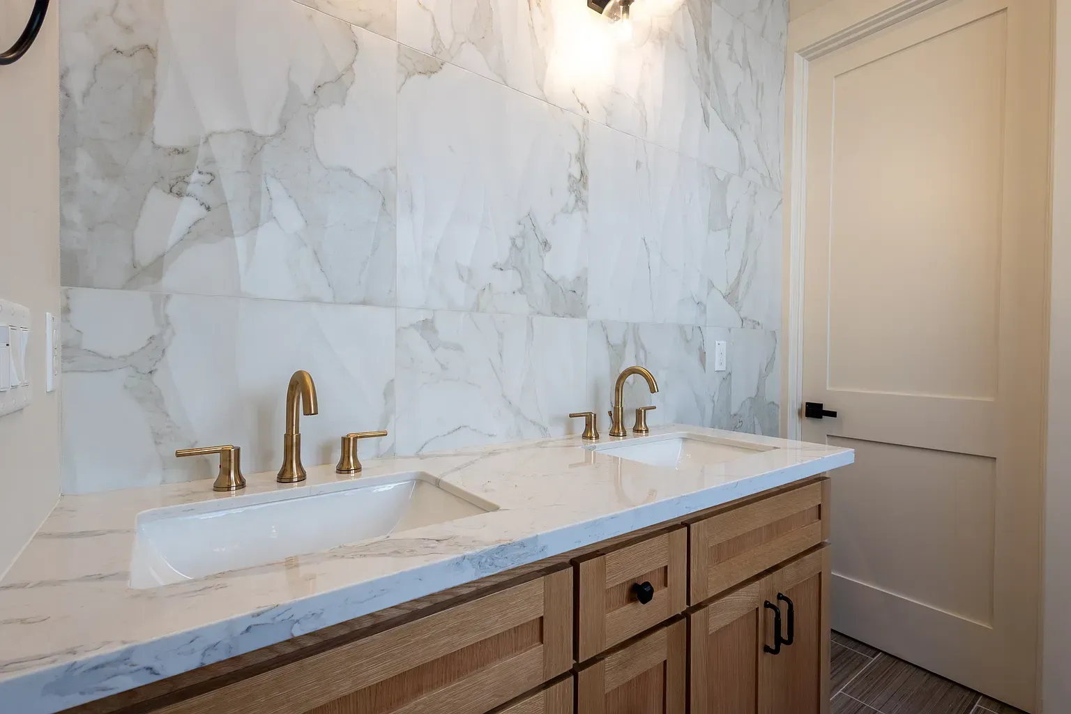 Bathroom with marble patterned wall, wood cabinets, white countertop, and gold faucets.
