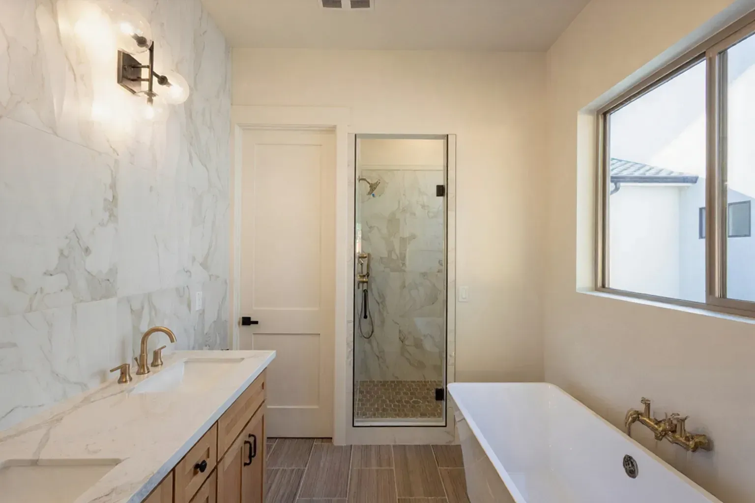 Bathroom with marble accent wall, glass shower, wooden vanity, and tub by a window.