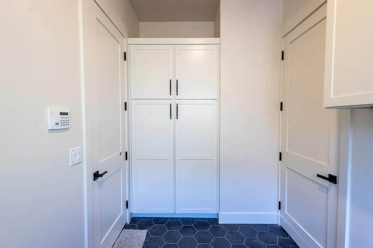 White pantry cabinets between two doors in a room with black hexagon tile flooring and off-white walls.
