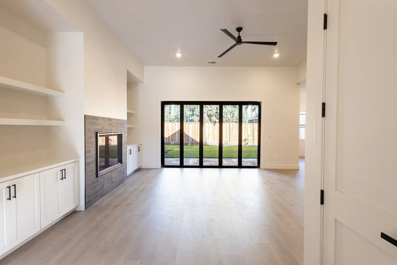 Bright, empty living room with built-in shelves, fireplace, and folding glass doors leading to a yard.