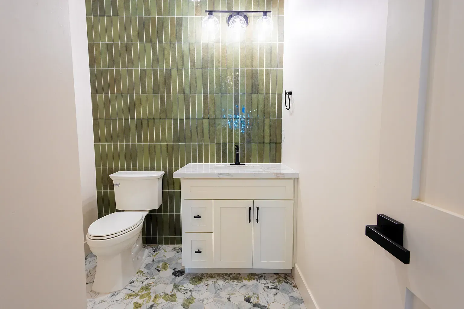 Small bathroom with green tiled accent wall, white vanity, and patterned floor.