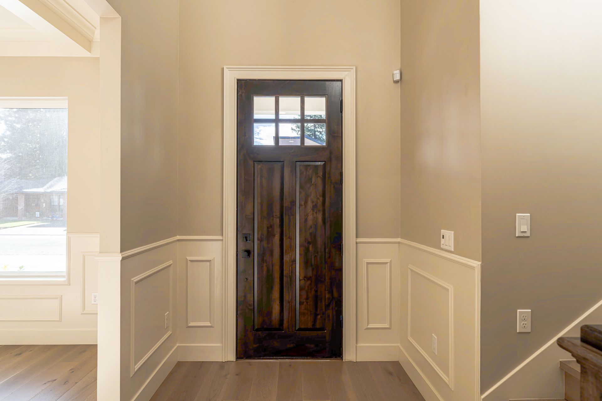 Wooden front door with glass panels, beige walls, and white wainscoting.