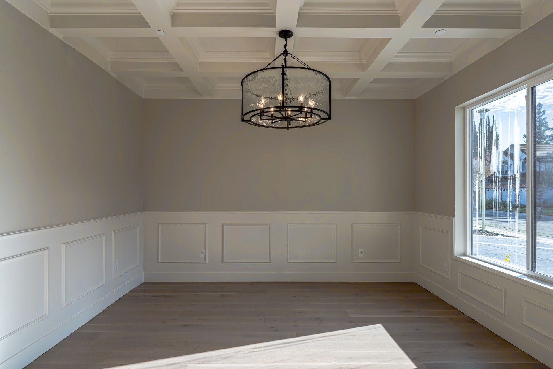 Empty dining room with crown molding, wainscoting, chandelier, and large window; light gray walls and wood floors.