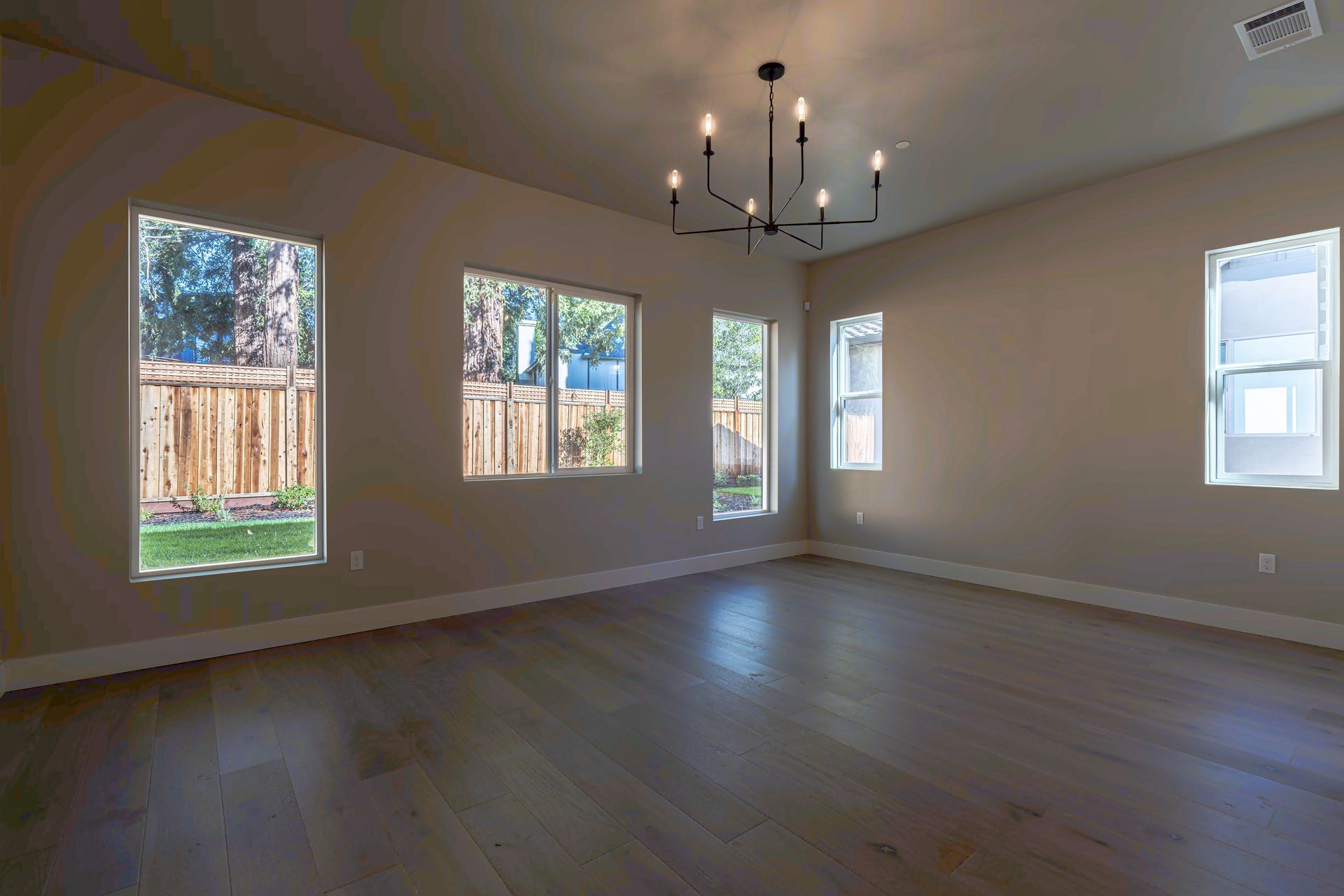 Empty room with wood floors and windows overlooking a backyard. Black chandelier hangs from the ceiling.