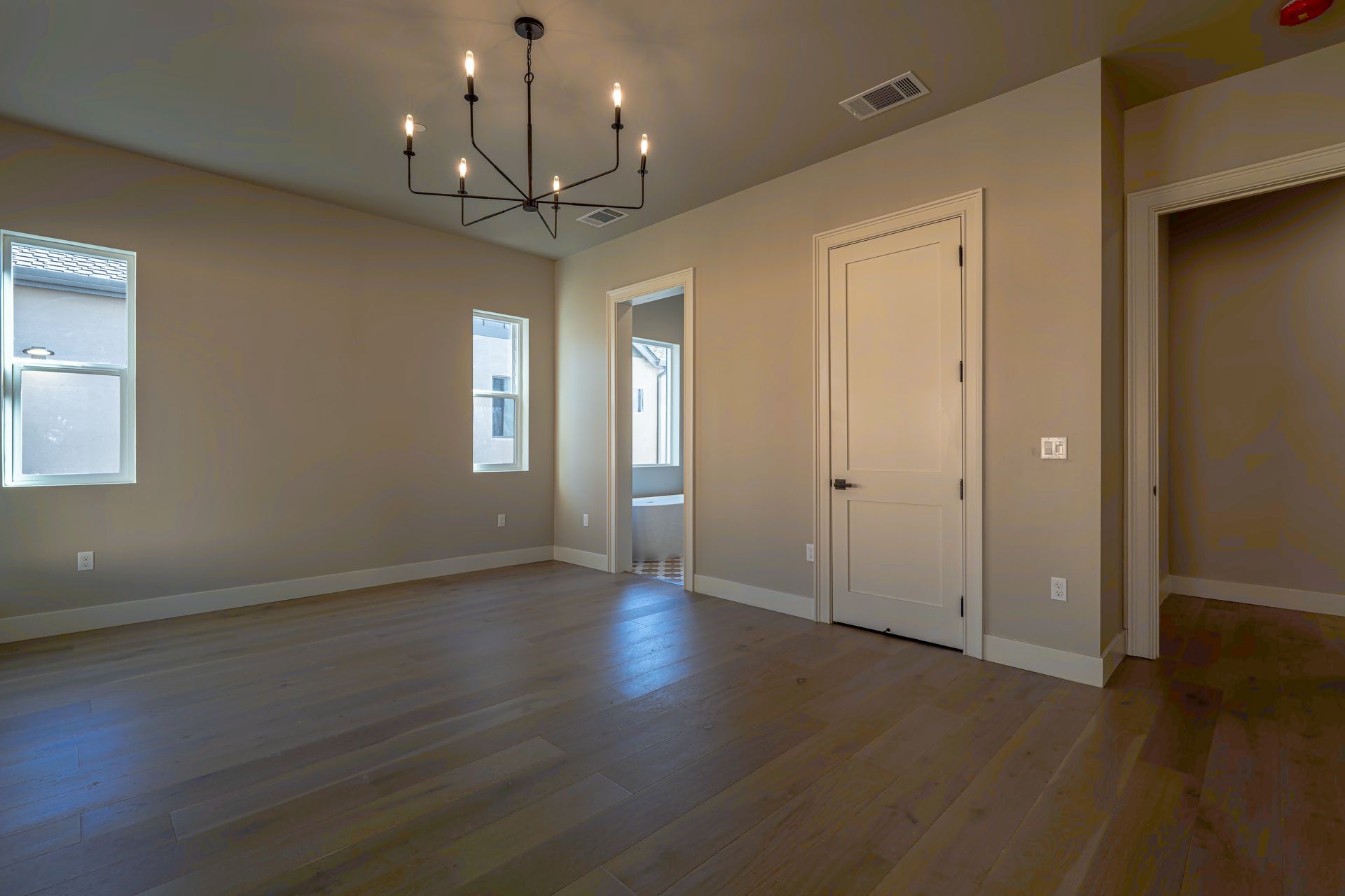 Empty room with wood floors, neutral walls, light fixture, windows, and a doorway.