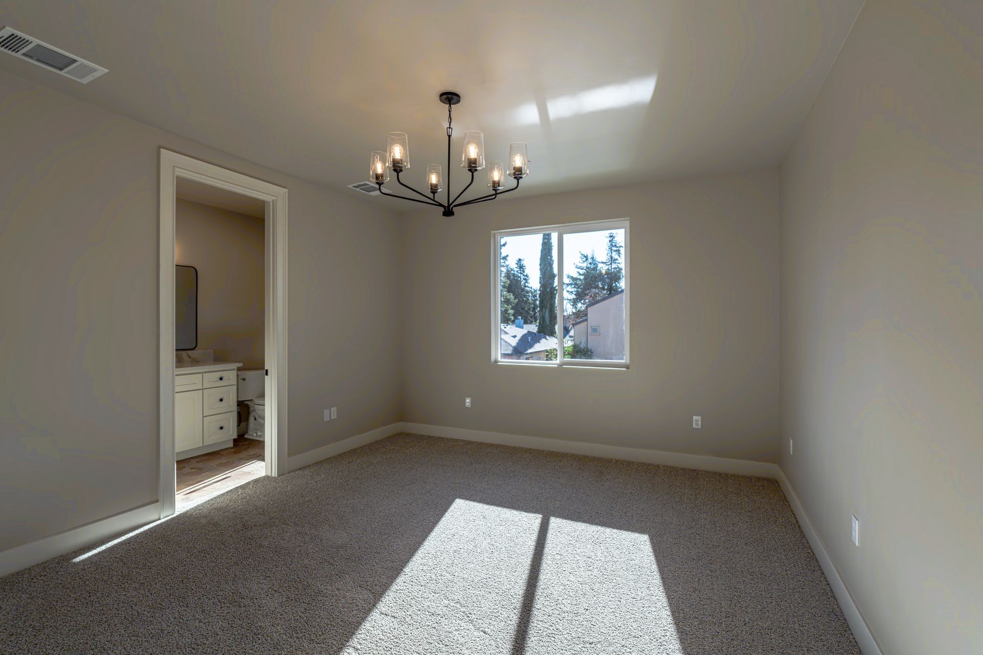 Empty room with a chandelier, window, and doorway to a bathroom with beige walls, and patterned carpet.