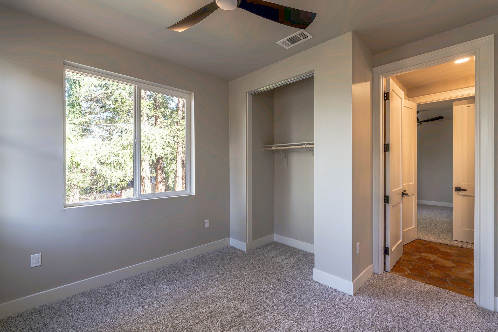 Empty bedroom with window, closet, and open doorway; light gray walls and carpet.