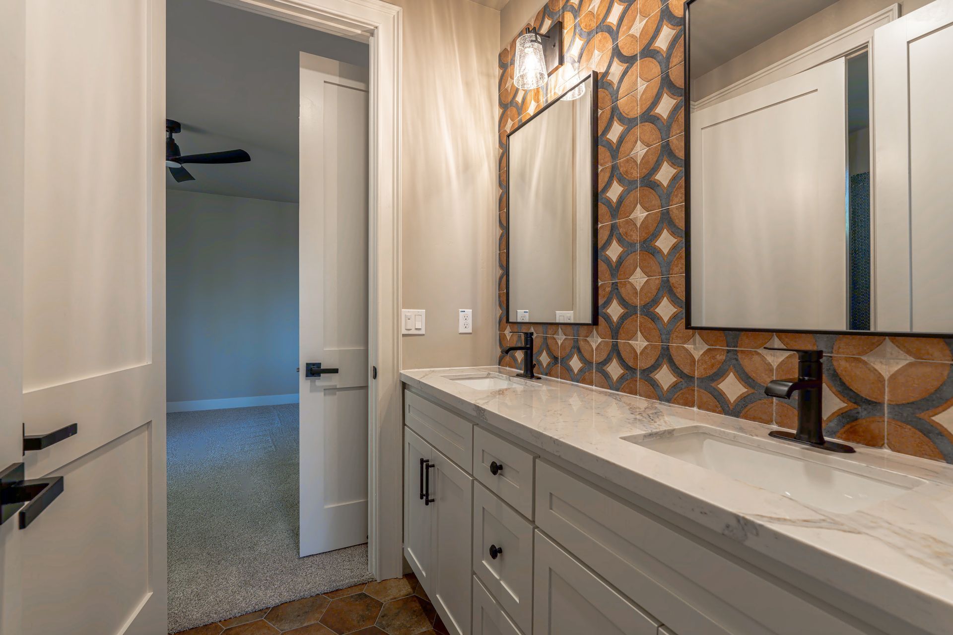 Bathroom with a double vanity and decorative tile backsplash; open door to bedroom.