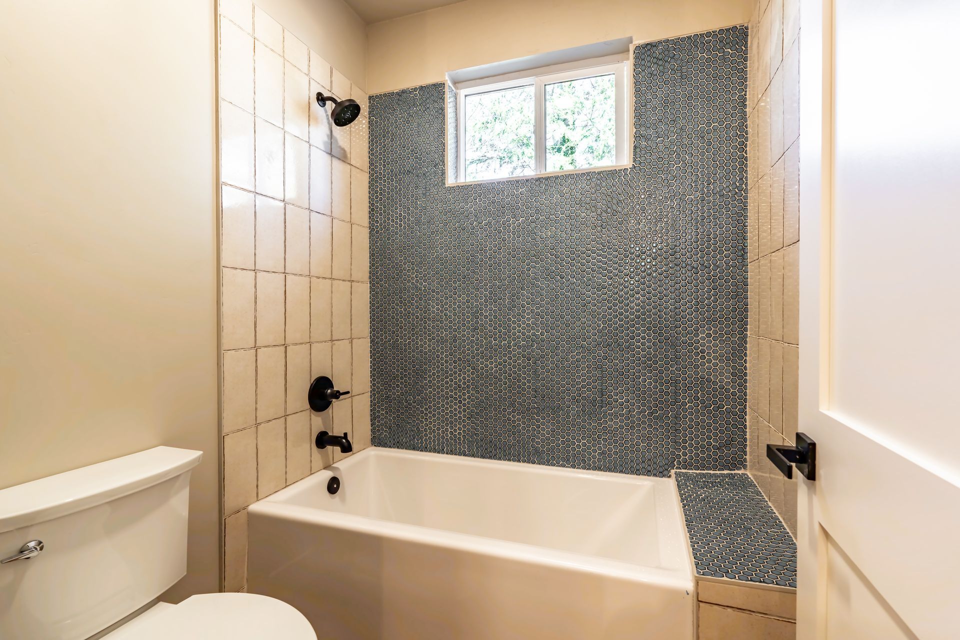 Bathroom with white tub, toilet, and light-colored walls. Gray tile backsplash and black fixtures.