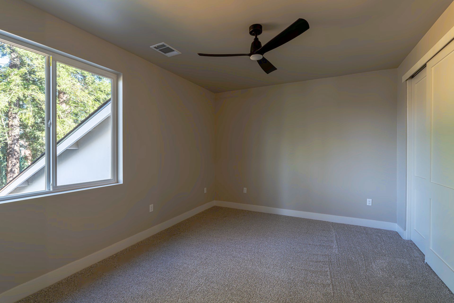 Empty bedroom with window, carpet, closet, ceiling fan, and beige walls.