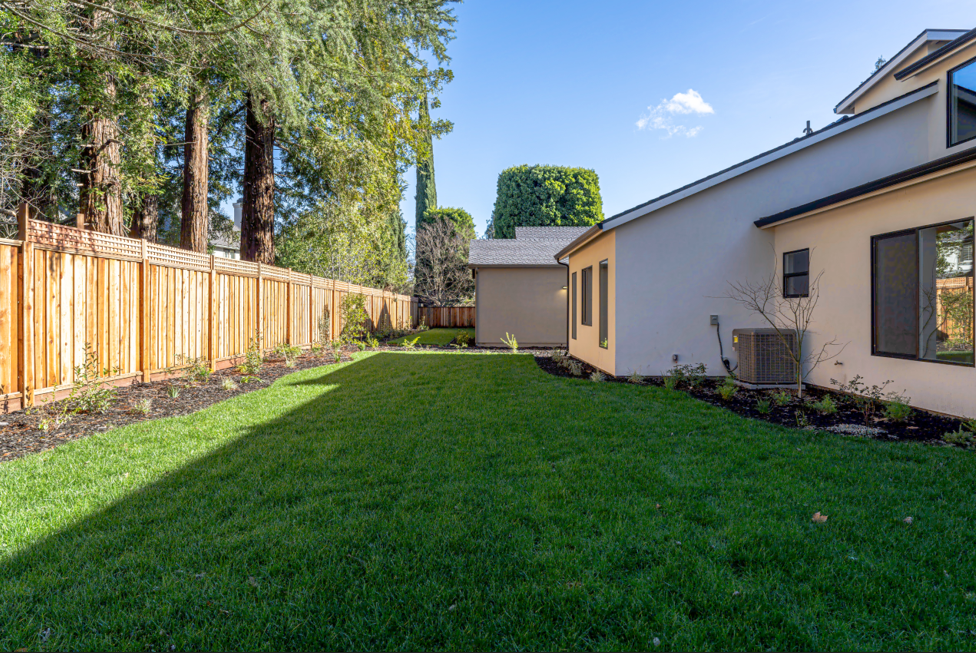 Backyard with green lawn, wooden fence, tan house, and trees under a blue sky.