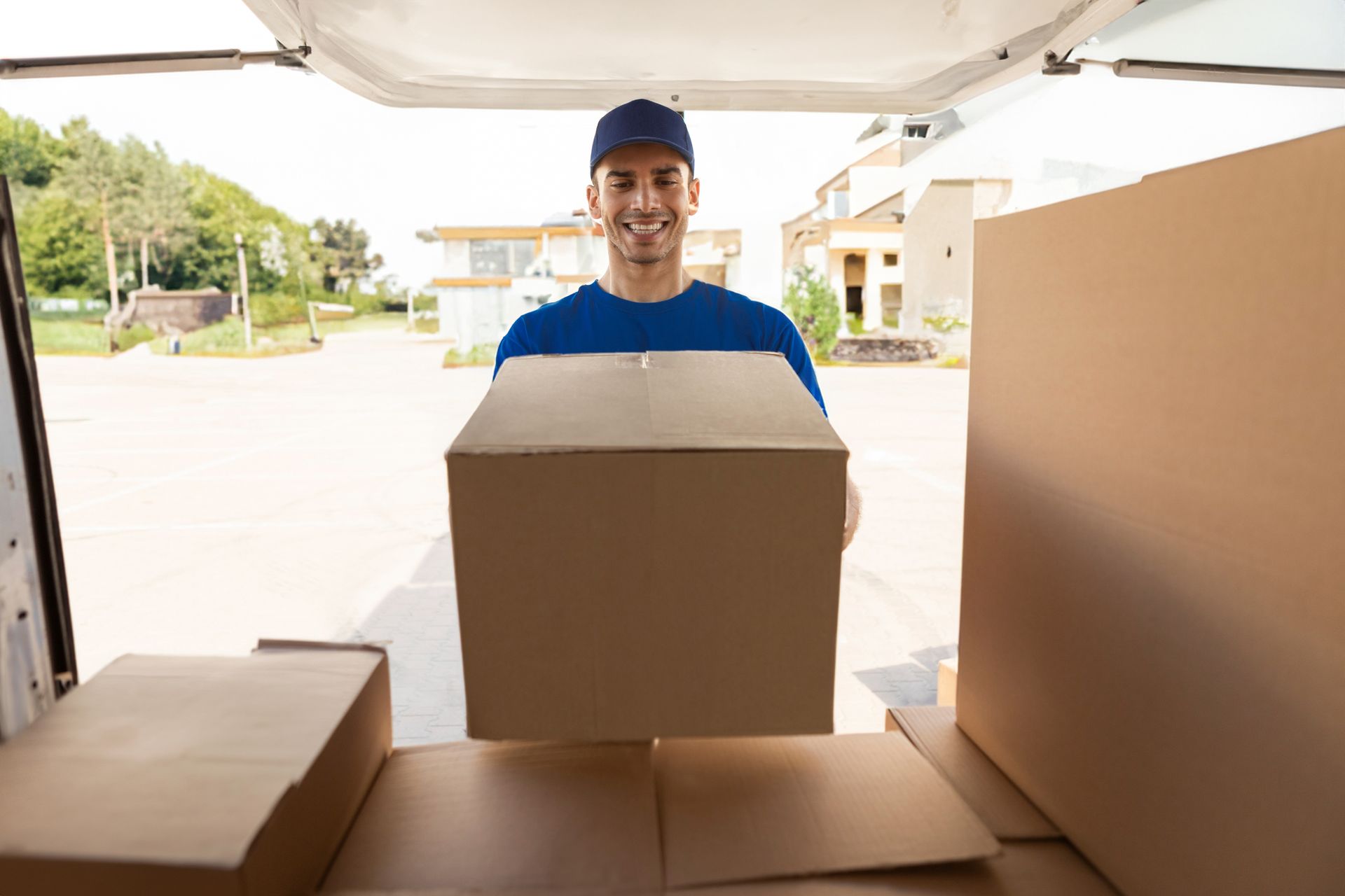 A delivery worker in a blue uniform smiling while holding a cardboard box in front of a delivery van.