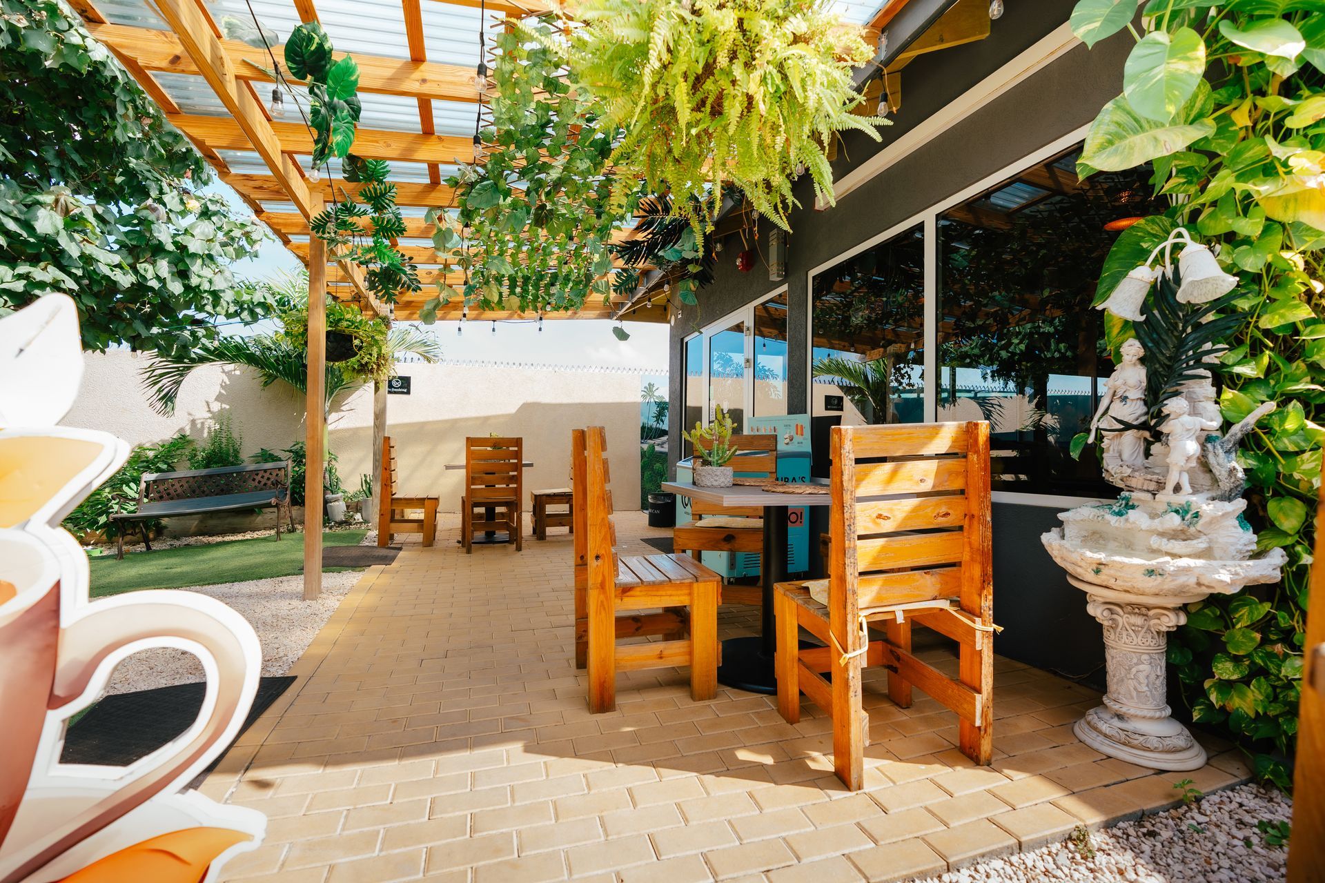 Outdoor patio with wooden tables, hanging plants, and a pergola beside a glass-walled building