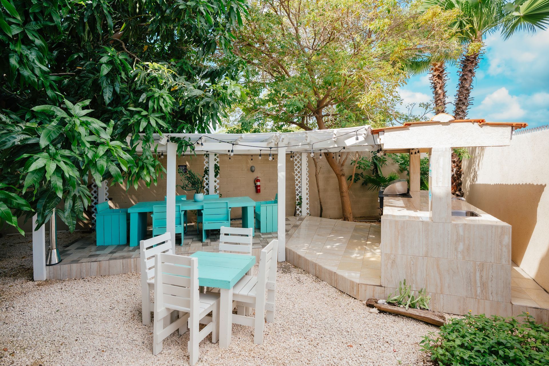 Outdoor patio with turquoise chairs and tables under a pergola, surrounded by trees and white walls