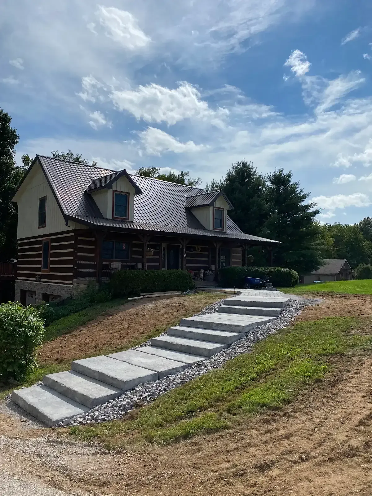 Log cabin with dormer windows and a stone walkway on a grassy slope under a partly cloudy sky