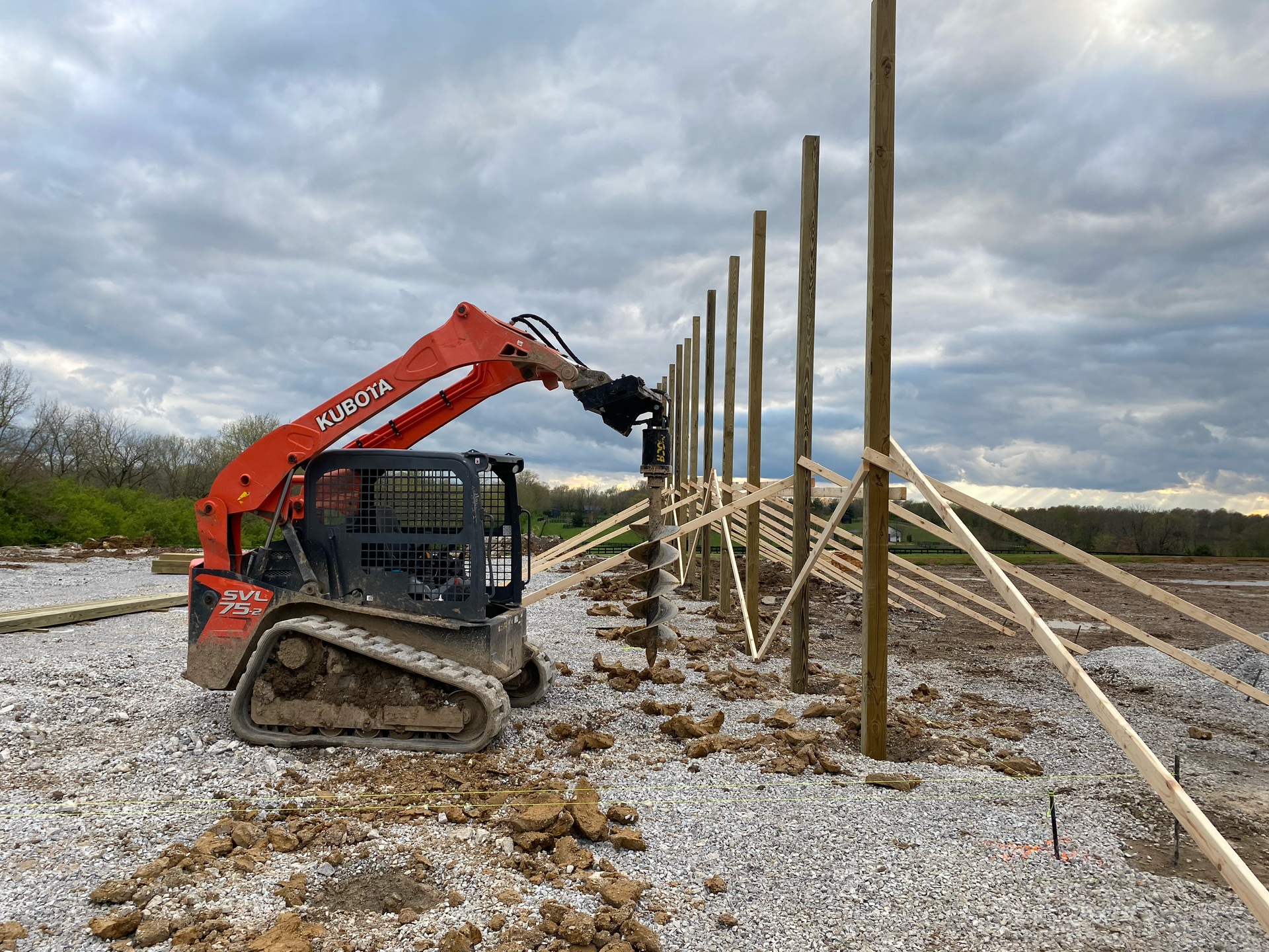 Wooden fence framework with vertical posts and diagonal braces in an open field under a cloudy sky