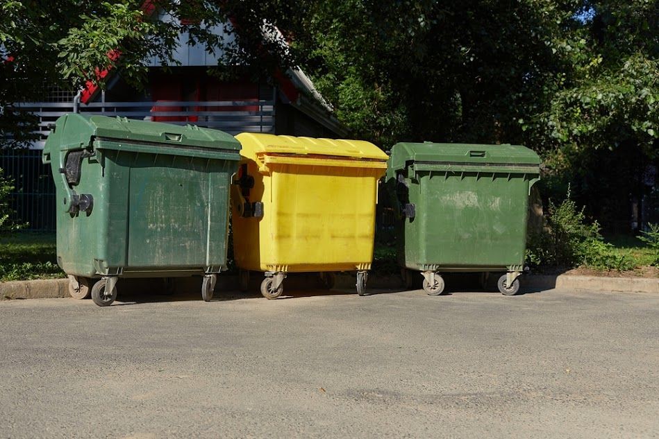 Three large recycling bins, green and yellow, sit on asphalt with trees in the background.