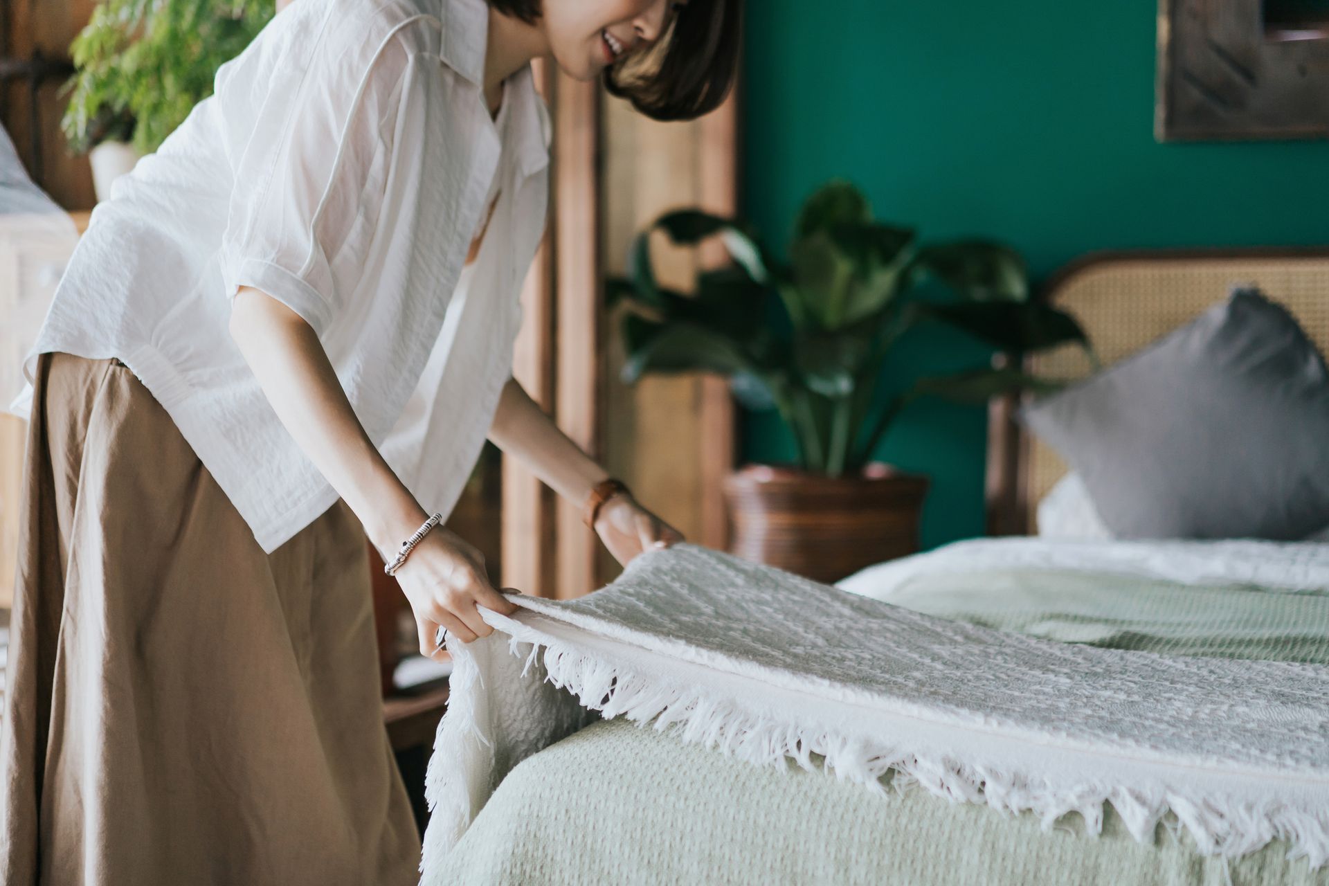 Woman in white shirt and tan pants making a bed in a teal bedroom with plants.