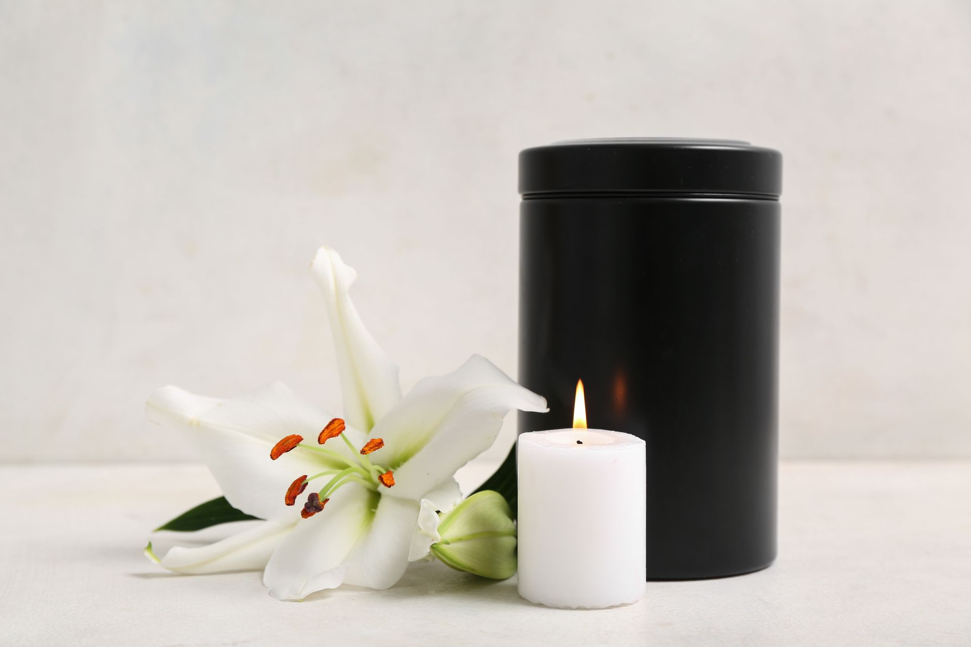 White lily and lit candle beside a black cylindrical urn on a light surface