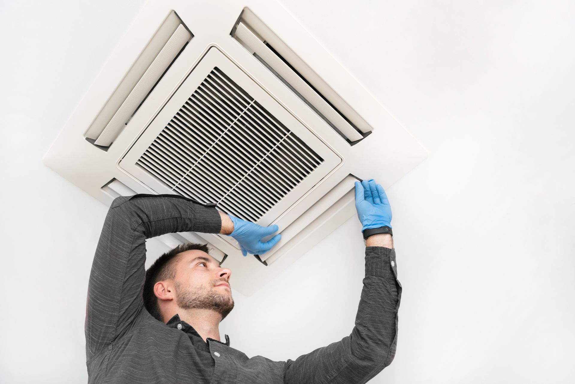 Man in blue gloves servicing a white ceiling air conditioning unit.